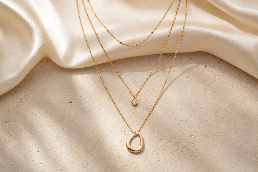 Three delicate gold necklaces arranged in graduated layers on ivory silk and stone